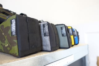 Straight Cut bike-packing bags in camouflage, grey, green, yellow and blue lined up on a shelf in the Edinburgh studio.