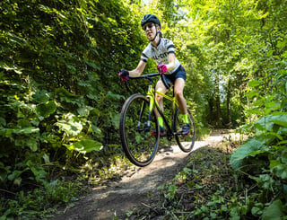 Rider in an All-City jersey hops the front wheel of a Macho King along a narrow wooded singletrack path.
