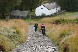 Two riders descend a rubble track towards whitewashed farm buildings in the Elenydd, Mid Wales.