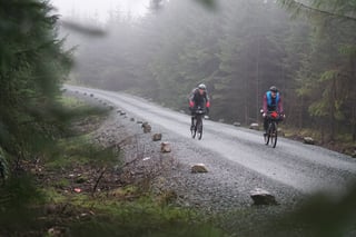 Two riders on a misty gravel forestry road through conifers on the Wildcat 100 route in Mid Wales.