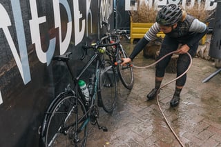 Rider in muddy kit rinses a road bike with a hose against a hoarding after the Backyard Breakaway.