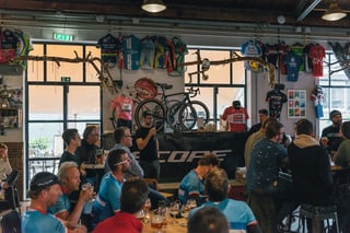 Speaker with a microphone in front of a Scope banner as riders drink beer at the Backyard Breakaway prize giving.