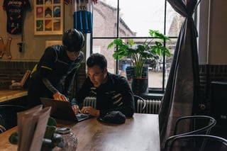 Two riders in cycling kit look at a laptop at a cafe table during the Backyard Breakaway in Eindhoven.