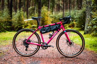 Salsa Journeyman gravel bike in hot pink with a frame bag and 650b tan-wall tyres, side view in a forest.