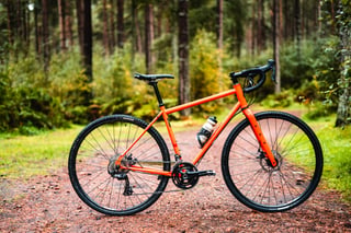 Orange steel Salsa Vaya gravel bike with drop bars and a bottle cage, side view on a forest track.