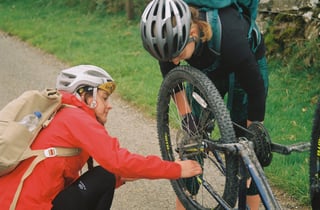 Two riders fix an upturned mountain bike at the roadside in the Lake District, checking the rear wheel and drivetrain.