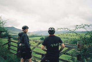 Two riders in helmets pause at a wooden gate, looking over green Lake District fells under a grey sky.