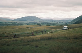 Riders on a grassy track across open Lake District fells, with a van parked on the hillside beyond.