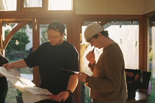 Two riders in caps study paper maps and checkpoint sheets in a sunlit room at the Low Key Lakes Weekender.
