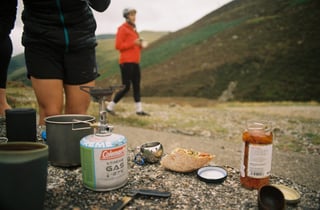 Camp stove, gas canister, pitta sandwich and jar of sauce on a rock while riders stand on the hillside behind.
