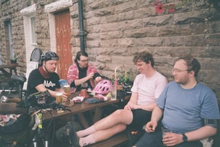 Four riders relax at an outdoor table by a stone cottage, with helmets, drinks and snacks in front of them.