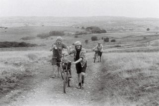 Two grinning riders push their bikes up a rough grassy track with a third riding behind, Zetland Cycles weekender.