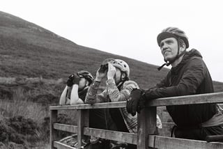 Three riders lean on a wooden fence below a fellside, one scanning the hills through binoculars, in black and white.