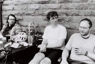 Three riders sit against a stone wall at a pub stop with drinks, crisps and a helmet on the table.