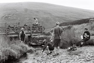 Riders rest and refill bottles at a stream by a small wooden footbridge, with bikes propped on the bridge.