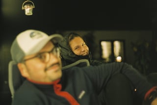 Two people in warm layers chat at night by a hanging candle lantern, one in the foreground out of focus.