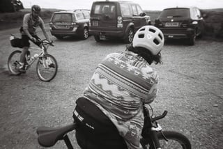 Black and white view from behind a rider in a patterned top as another rider cycles past parked cars at a car park.