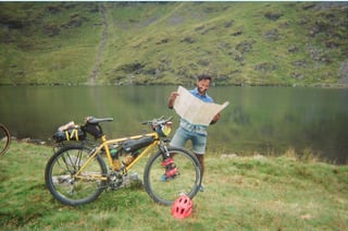 Rider reads a paper map beside a loaded yellow mountain bike on the shore of a Lake District tarn.