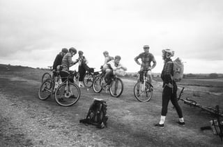 Group of riders and bikes gathered on a gravel patch for a break, a rucksack on the ground, in black and white.