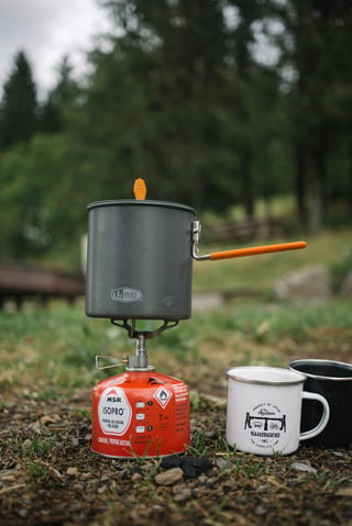 GSI pot on a stove screwed to an MSR IsoPro gas canister, with Shimano and black enamel mugs beside it on gravel.