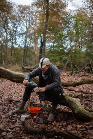Rider in a cap sits on a fallen tree in autumn woodland, pouring a drink into a mug beside a camp stove.