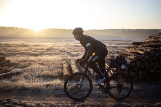 Rider on a gravel bike with a saddle bag passes log piles on a frosty heath at sunrise on a daypacking ride.