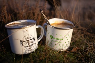 Two enamel mugs of coffee, one Shimano and one komoot, sit in the grass in low sun on a daypacking ride.