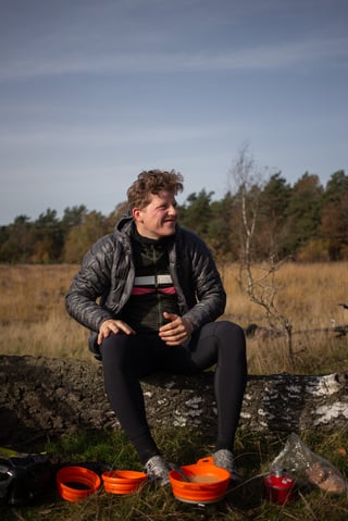 Smiling cyclist in a down jacket sits on a fallen birch trunk beside orange bowls and a gas canister on Dutch heathland.