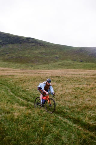 Rider in a striped shirt and sunglasses on an orange gravel bike crosses rough grass below a Lake District fell.