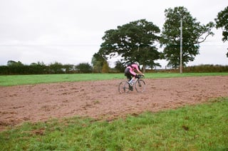 Rider in a pink and black kit crosses a ploughed field on a road bike, with large trees behind.
