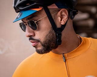 Close-up of a rider in an orange Attacus Cycling jersey, blue cycling cap under a black helmet, and sunglasses.