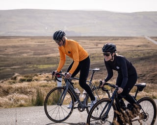 Two riders on road bikes ride along a moorland road in Attacus Cycling kit, one in an orange long-sleeve jersey.