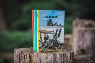 Four copies of the Bikepacking Journal standing on a tree stump, the front cover showing a loaded bike against a white shed.