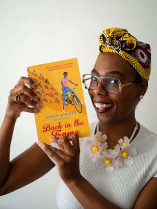 Jools Walker holds up her book Back in the Frame, smiling in a yellow headwrap and glasses.
