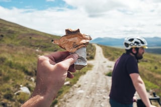 Hand holding a half-eaten Outdoor Provisions Bara Brith energy bar on a gravel track with a rider in the background.