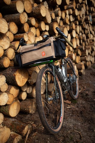 Detour Studio tan and black handlebar bag on the front rack of a gravel bike parked against a stack of logs.