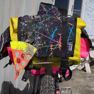 Wizard Works saddle bag in black splatter fabric with yellow body and pink pockets, muddy on a bike rack.