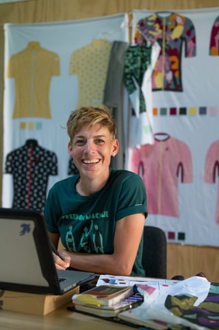 Iris Slappendel smiling at her desk in front of I-ris.cc cycling jersey designs pinned to the wall.