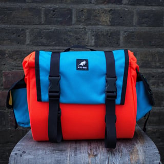 Wizard Works saddle bag in orange with a bright blue flap and side pockets, on a stool in front of a brick wall.
