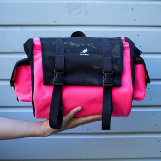 Wizard Works saddle bag with pink body and black camouflage flap, held up against a pale blue wall.