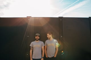 Two Outdoor Provisions founders in matching grey logo T-shirts laughing in front of a dark wall in low sun.
