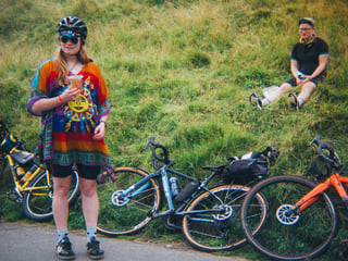 Low Key Lakes Weekender rider in a colourful poncho holds a drink beside bikes lying on a grassy verge.