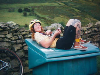 Rider in a yellow helmet lies back on a green grit bin beside a drystone wall in the Lake District, film camera in hand.