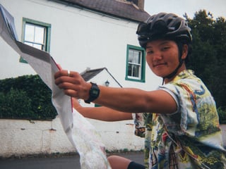 Rider in a patterned shirt and helmet holds up a paper map in a village on the Low Key Lakes Weekender.