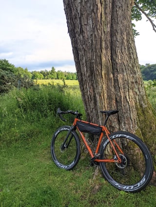 Orange Fairlight Secan with Scope R4A wheels leans against a tree beside a field on a summer evening.