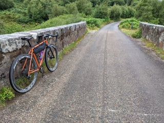 Orange Fairlight with Scope R4A wheels leans on a stone bridge wall on a quiet road near Rhiconich.