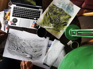 Pen and watercolour drawings of the Stelvio Pass hairpins on a desk beside a laptop, Stef's Stelvio sketches in progress.