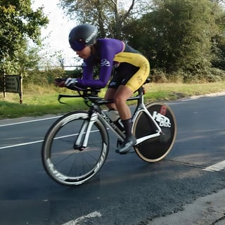 Rider in purple and yellow kit on a time trial bike with a disc rear wheel, riding along a country road.