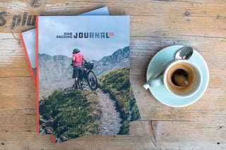 Bikepacking Journal issue 01 on a wooden table beside an espresso, cover showing a rider with a bike on a mountain trail.