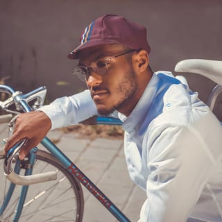 A man in a maroon striped Vera cycling cap and round glasses rests an arm on a blue vintage road bike.